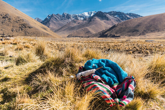 Andean blanket and spinning wheel with alpaca fiber lying in the field surrounded by snowy mountains and blue sky with green and yellow vegetation