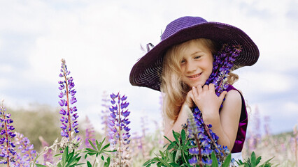 Shy girl in a hat holds a lupine in a field of flowers.