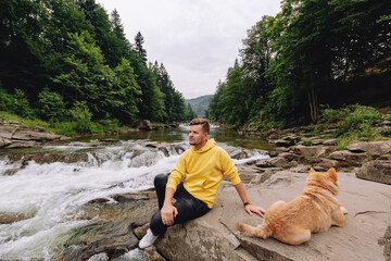 A man near a mountain river while sitting on a rock. A brown dog is running nearby.