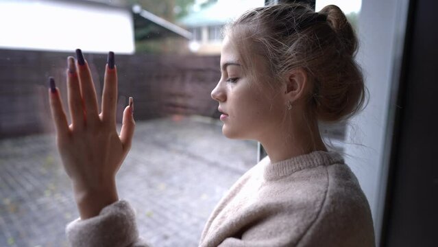 Side view portrait of sad brokenhearted teenage girl looking out the window on rainy day touching glass. Depressed frustrated Caucasian beautiful teenager thinking standing indoors at home