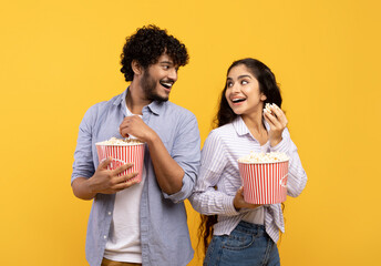 Overjoyed indian man and woman eating popcorn, standing back to back and smiling at each other,...