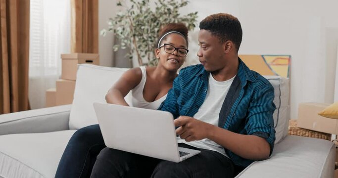 A married couple sits on the couch in their new apartment after moving in, the man holds laptop in his lap, they search together for furniture, furnishings, decorations on websites