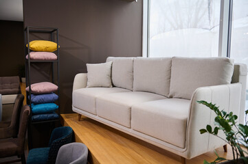 Stylish minimalistic white sofa with pillows displayed for sale in a showroom of modern furniture, next to stand with colorful cushions from fabrics of different texture, softness, colors and quality