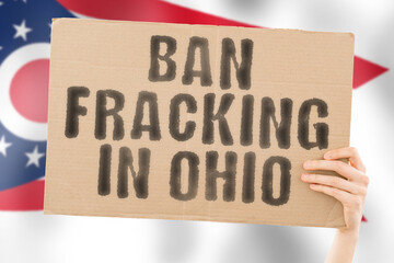 The phrase " Ban fracking in Ohio " on a banner in men's hand with blurred Ohio flag on the background. Nature. Extract. Stop. Pipe. Energy. Drill. Raw. Power. Fuel. Fossil. Crisis