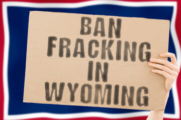 The phrase " Ban fracking in Wyoming " on a banner in men's hand with blurred Wyoming flag on the background. Nature. Extract. Stop. Pipe. Energy. Drill. Raw. Power. Fuel. Fossil. Crisis