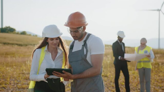 International team of business partners standing together among wind turbines and examining blueprints. Group of professionals cooperating for producing clean eco friendly energy. Wind turbines.