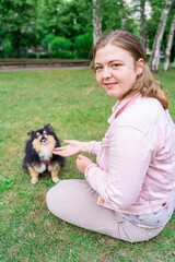 Young blond woman in pink clothes training and feeding her fluffy small black dog pet Spitz Pomeranian, sitting in summer park or backyard on green grass loan near flowers and trees. Friendship