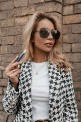 Stylish beautiful young woman with sun glasses smokes an e-cigarette and walks in the city near a brick wall