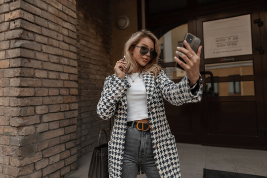 stylish beautiful blond girl in a fashionable coat with glasses and a bag walks in the city and takes a selfie photo on a smartphone