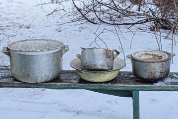 a row of old dirty metal bowls and pans stand on a wooden bench in white snow on a winter street