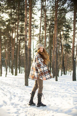 A beautiful young girl with green eyes in a hat and a plaid shirt is spinning and standing on a winter snowy day in a forest city park. Beautiful background and fashionable clothes, healthy clean skin