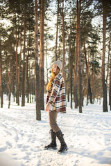 Fototapeta premium A beautiful young girl with green eyes in a hat and a plaid shirt is spinning and standing on a winter snowy day in a forest city park. Beautiful background and fashionable clothes, healthy clean skin