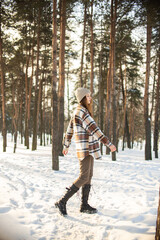 A beautiful young girl with green eyes in a hat and a plaid shirt is spinning and standing on a winter snowy day in a forest city park. Beautiful background and fashionable clothes, healthy clean skin