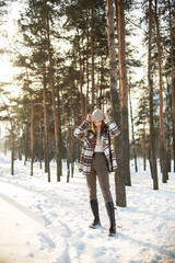 A beautiful young girl with green eyes in a hat and a plaid shirt is spinning and standing on a winter snowy day in a forest city park. Beautiful background and fashionable clothes, healthy clean skin