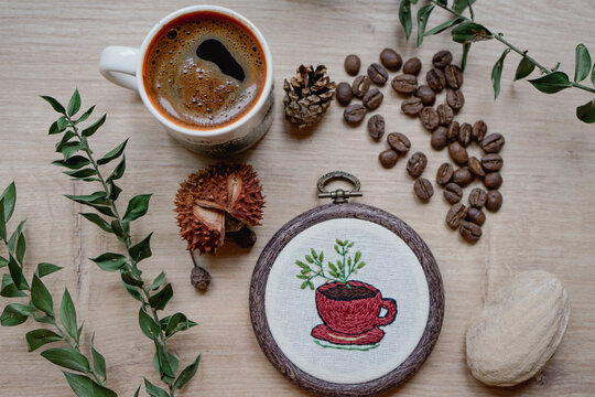 Coffee mug embroidery with hot coffee and coffee beans on wooden background, top view, flat desk. Hand made embroidery, new hobby concept for new year.