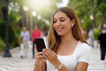 Excited young woman watching her smartphone when walking in the street with blurred people on the background. Beautiful girl using mobile app outdoors. Teenager lifestyle technology concept. - obrazy, fototapety, plakaty
