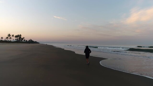 A man is busy with a morning jog at a slow pace along the seashore on the beach next to the ocean at dawn. Sports training, running along the surf line. A man in shorts, hoodies and headphones runs.