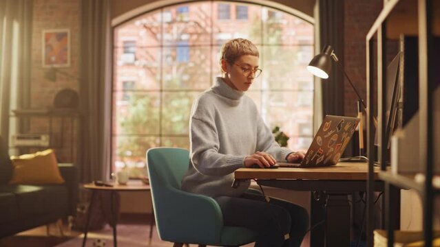Young Diverse Woman Working from Home on Laptop Computer with Stickers in Sunny Stylish Loft Apartment. Creative Designer Wearing Cozy Blue Sweater and Glasses. Urban City View from Big Window.