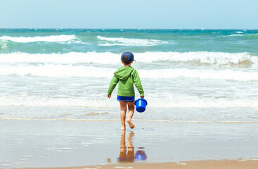 Little boy alone on the beach walking towards the water with a plastic bucket in his hand.