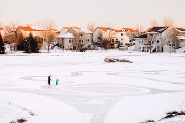 A father and daughter ice-skate on a neighborhood pond surrounded by houses