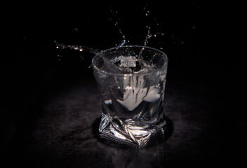Splashes of water from a piece of ice falling into a transparent glass with a black background