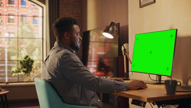 Young African American Man Working from Home on Desktop Computer with Green Screen Mock Up Display. Creative Male Checking Social Media, Browsing Internet. Urban City View from Big Window.