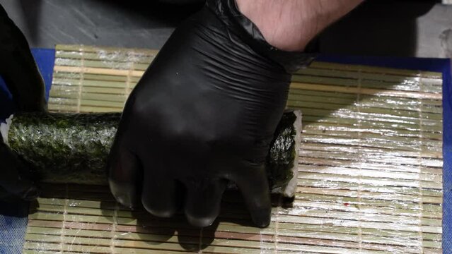chef in kitchen in black sterile gloves spins long suchi roll in bamboo mat on blue board