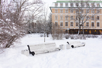 Snow-covered benches in winter park with bare snowy trees and bushes against five-story building