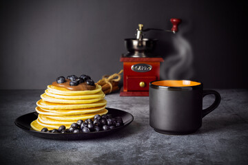 Pancakes with blueberries and chocolate paste on black dish, cup of coffee with steam, coffee grinder and cinnamon sticks on gray table on black background