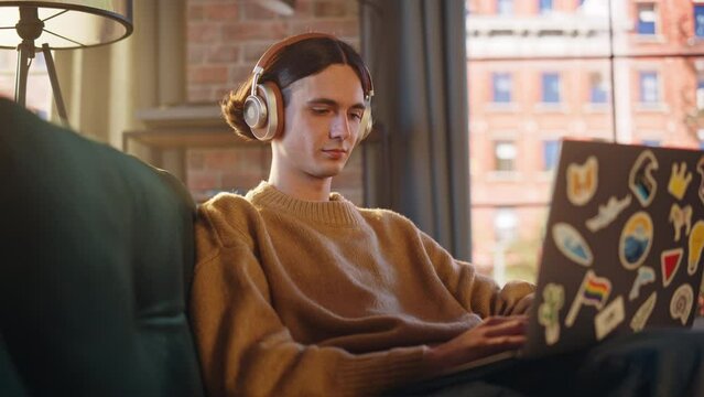 Young Handsome Man Sitting on Sofa and Using Laptop Computer in Sunny Stylish Loft Apartment. Creative Designer Wearing Cozy Yellow Sweater and Headphones. Urban City View from Big Window.