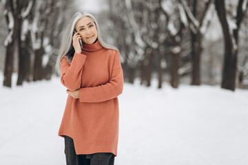 Portrait of senior woman with gray hair in knitted sweater talking by phone on background of snowy weather in winter