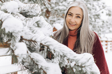 Senior woman with gray hair in a knitted sweater and scarf, sincerely smiling holding a Christmas...