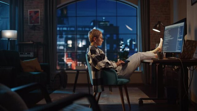 Young Woman Writing Code on Computer in Stylish Loft Apartment in the Evening. Creative Female Wearing Cozy Clothes, Resting Feet on Table and Using Wireless Keyboard. Urban City View from Big Window.