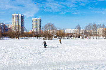 Winter ice fishing, lake, frosty day. A fisherman is engaged in ice fishing in the pond of the city park. Against the background of a multi-storey, modern, residential development.