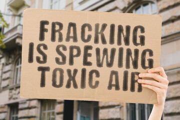 The phrase " Fracking Is Spewing Toxic Air " on a banner in men's hand with blurred background. Disease. Issue. Dirty. Crude. Pump. Fire. Drill. Cloud. Air. Rig. Oil. Shale. Gas. Fuel. Dusk. Pollution