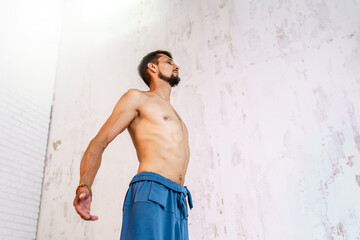 A bearded man is naked to the waist in sweatpants against a white wall. Muscular strong man doing breathing practice