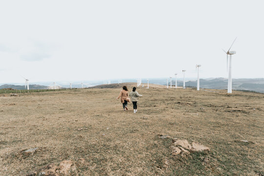 Vintage cinematic image of two young woman running between electrical wind mills with soft tones. Spring ambient freedom and liberty concepts. Friendship and road trip vibes - Powered by Adobe