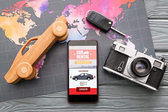 Mobile phone with open car rent app, toy, photo camera and map on dark wooden background