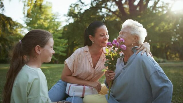 Small girl with mother giving cake and flowers to grandmother in a park, celebrating birthday.