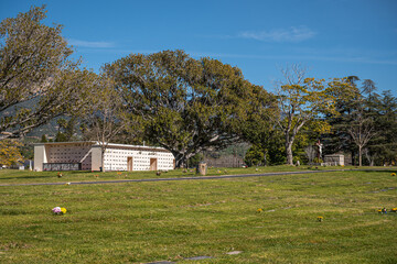Obraz premium Santa Barbara, California, USA - February 8, 2022: Calvary Cemetery. Landscape of green burial lawn in front of white burial building. Plenty of trees, all under blue sky.