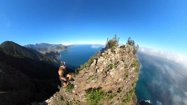 A young and strong solo hiker is walking along the steep edge of Espigao Amorelo in Madeira with a 360 action camera on a selfie stick with an amazing view of the landscape and ocean beneath him.