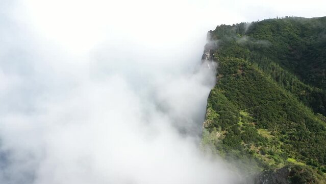 Drone shot of Espigao Amorelo Cliff Side in Madeira Portugal. Surrounded by clouds with lots of greenery from trees on the side. Dramatic and epic mountain with clean and fresh air.