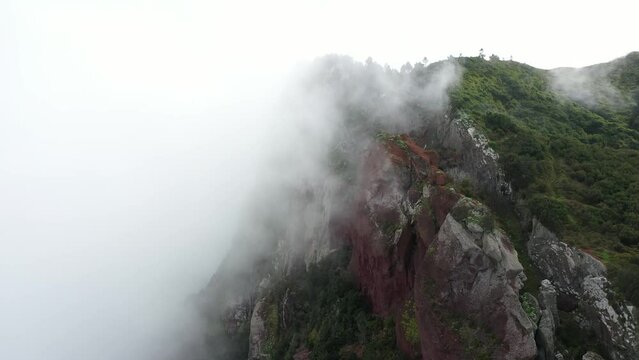 Drone shot moving up and back, revealing the cliffside of Espigao Amorelo in Madeira with thick clouds moving and surrounding the peak. Lush and green forest on the steep side.