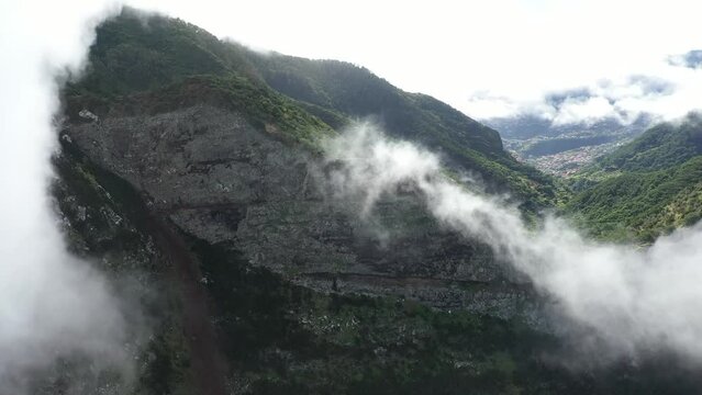 Drone shot moving up tilting down, revealing the cliffside of Espigao Amorelo in Madeira with thick clouds moving and surrounding the peak with a village in the distance.