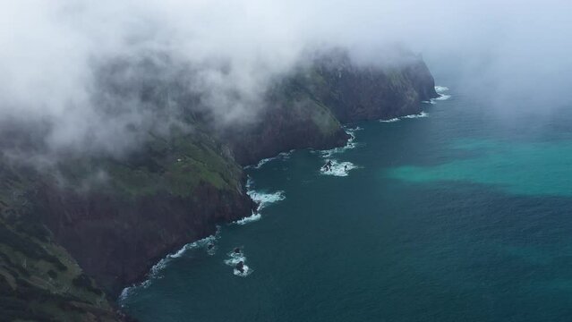 Drone shot of the cliff side of Espigao Amorelo in Madeira coming down out of the clouds and reveals the dramatic and epic mountain with the turquoise and blue ocean below it.