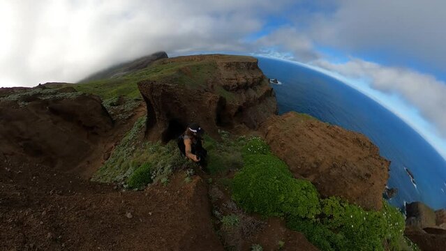 Solo hiker walking up Espigao Amorelo mountain in Madeira with a 360 action camera. Beautiful green cliffside with some clouds and blue sky. His friend is waiting at the top.