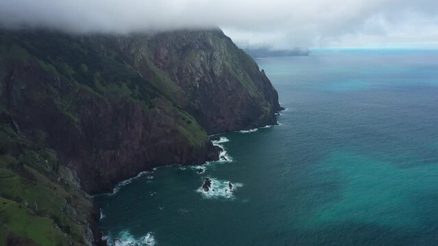 Drone shot of the cliff side of Espigao Amorelo in Madeira with its dramatic and steep cliff. Dark blue and turquoise water with reefs below as the waves crashes against the rocks. Thick clouds above.