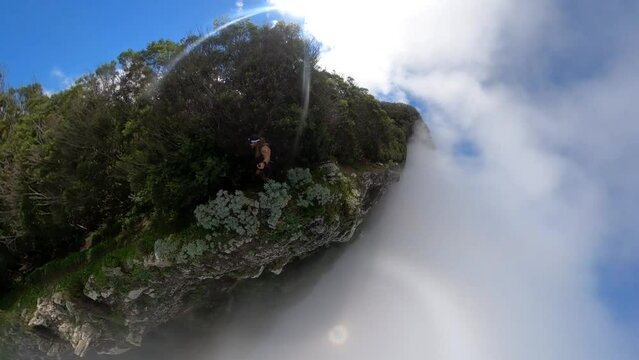 A young and strong solo hiker is running along the steep edge of Espigao Amorelo in Madeira with thin clouds around him during a sunny day with clear blue sky.