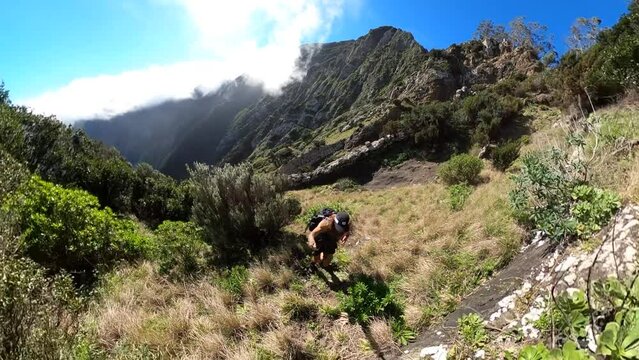 A young and strong solo man hiker is holding a selfie stick with a 360 action camera as he walks up the mountainside of Espigao Amorelo mountain in Madeira with the harch sun during a clear day.