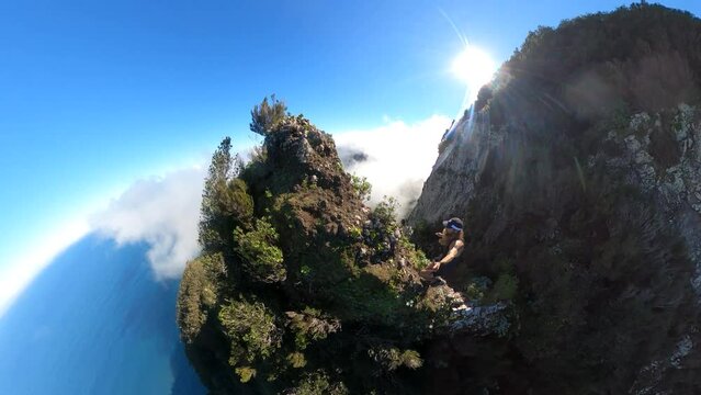 A young and strong solo hiker is walking along the steep edge of Espigao Amorelo in Madeira with thin clouds around him as he takes a dangerous step up to a rock on the cliffside to enjoy the view.